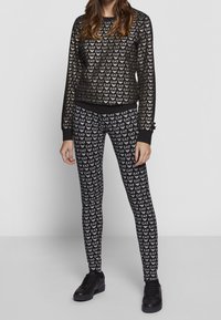 Černý vzorovaný outfit skládající se z dlouhého rukávu a legín zdobených opakujícím se designem loga, doplněný černými teniskami.