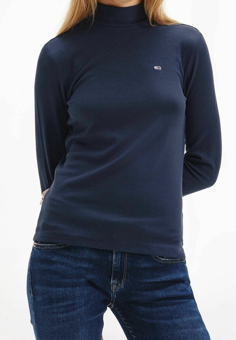 Marineblauwe geribbelde turtleneck shirt met lange mouwen en een klein logo op de borst. Gecombineerd met donkerblauwe jeans. Zachte textuur.