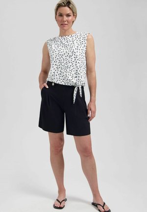Vrouw staat en draagt een witte mouwloze top met zwarte halvemaanpatronen en zwarte knielange shorts met zwarte sandalen.
