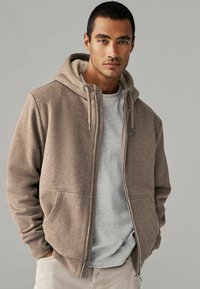 Rudas zip-up džemperis su pūkinėmis kapisonu, dviem priekiniais kišenėmis ir reguliuojamais raišteliais. Dėvimas ant šviesiai pilkos marškinėlių.