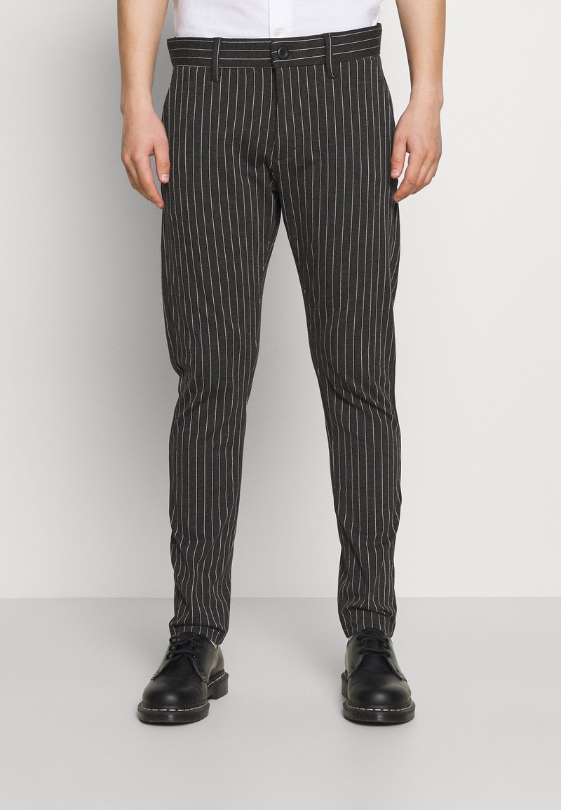 Homme portant un pantalon noir à fines rayures et des chaussures en cuir noir, debout devant un fond clair uni.