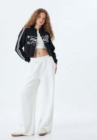 Giacca nera corta con strisce bianche e stampa grafica; pantaloni bianchi a gamba larga con vita elasticizzata; scarpe chiare con suola testurizzata.
