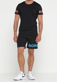 Svart kortärmad t-shirt med korallfärgat "BORG"-tryck på ärmarna, matchad med svarta shorts som har en stor blå "BORG"-logotyp, och vita sneakers.