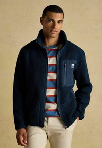 Nuori mies, jolla on päällä tummansininen fleece-takki sinivalkoraidallisen paidan ja vaalean beigepunaiset housut, seisoo käsi taskussa.