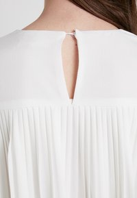 Robe blanche avec une texture plissée, dotée d'une petite ouverture en trou de serrure à l'arrière, fermée par un bouton.