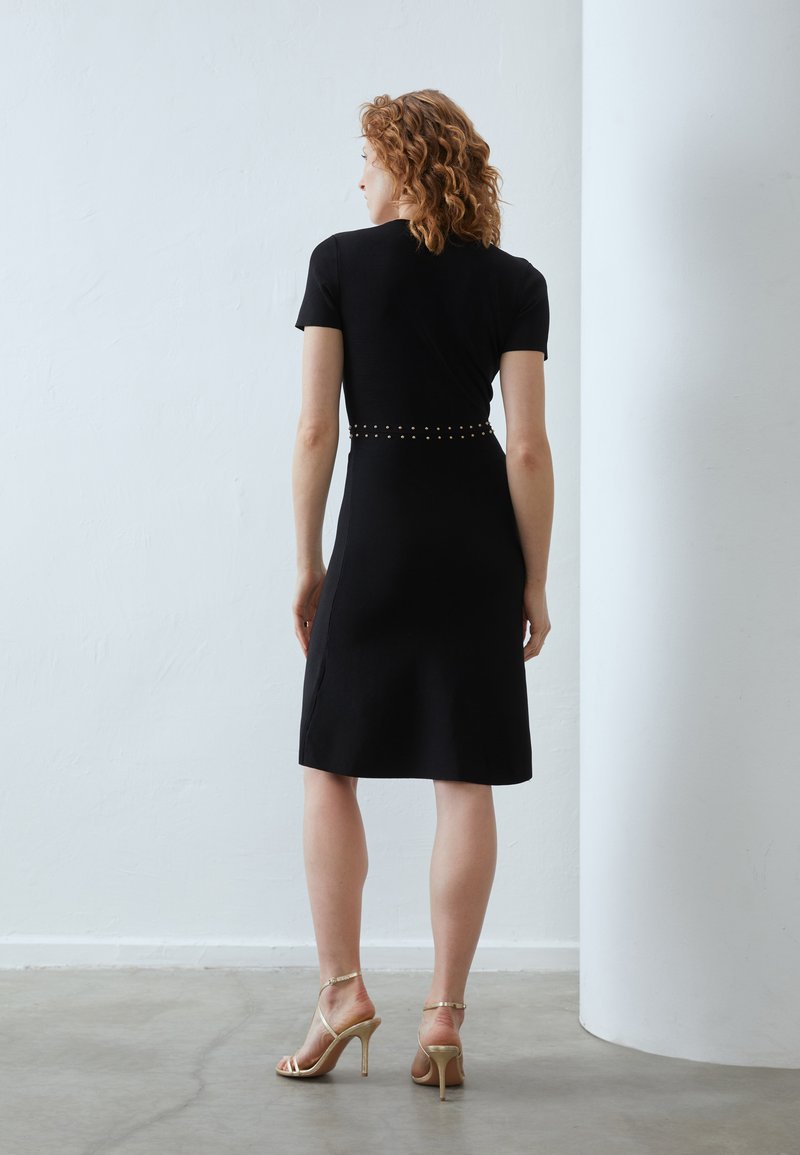 Czarna dopasowana sukienka z krótkimi rękawami, ozdobiona złotymi ćwiekami wzdłuż talii. Gładki materiał, do kolan, w połączeniu ze złotymi sandałami na obcasie.