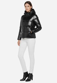 Giacca a vento nera con finitura lucida, cappuccio, chiusura frontale con zip e due tasche laterali. Indossata con pantaloni bianchi e stivaletti neri.