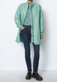 Světlounce zelená prošívaná bomber bunda s žebrovanými manžetami, nošená přes vzorovanou košili a tmavé úzké džíny; černé boty na platformě doplňují vzhled.