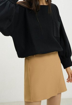 Vrouw draagt een zwarte top met lange mouwen en een zak, en een beige rok tot op de knie, staand met een arm licht omhoog.