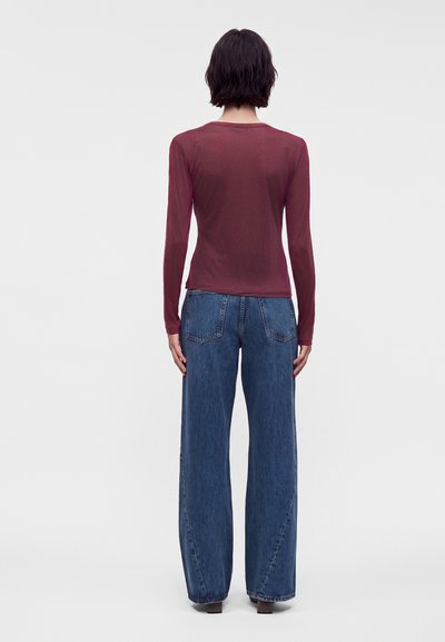 Personne aux cheveux courts et foncés, de dos, portant un haut bordeaux moulant à manches longues et un jean bleu foncé ample sur un fond uni.