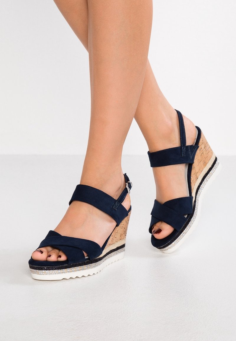 Jambes portant des sandales compensées bleu marine avec talons en liège et brides à la cheville, debout sur un sol blanc lisse.