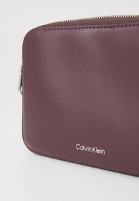 Bordeaux leren ritsportemonnee met een gladde textuur en zilveren accenten, met het "Calvin Klein" logo in wit gebosseleerd.