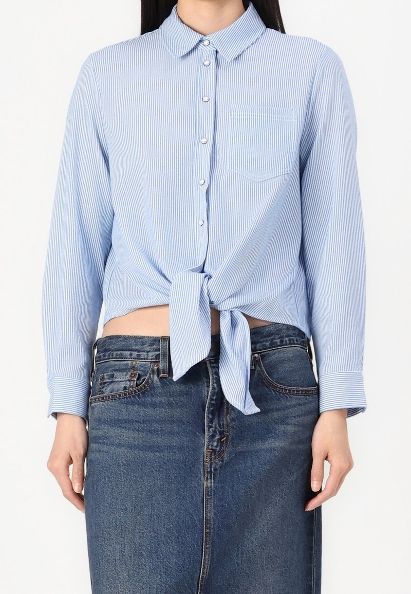 Femme portant une chemise rayée bleu clair et blanc, boutonnée, nouée à la taille avec une poche devant, associée à une jupe en jean taille haute.