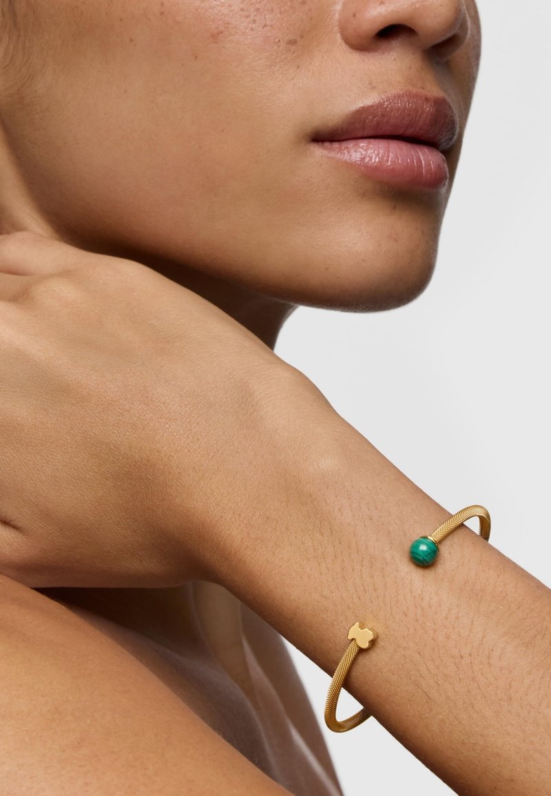 Primer plano de la muñeca de una mujer que lleva una pulsera de oro con una piedra verde y un pequeño colgante de mariposa dorada.