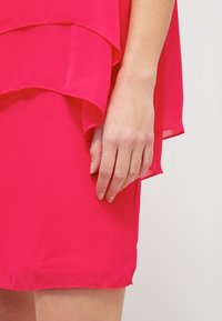 Dłoń kobiety spoczywająca obok jasnoróżowej, warstwowej spódnicy na jasnym tle, ukazująca fakturę tkaniny i naturalny odcień skóry.