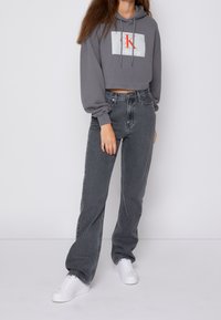 Sudadera gris corta con un logo rojo y blanco, combinada con jeans oscuros de tiro alto y zapatillas blancas. Material de algodón suave, corte relajado.