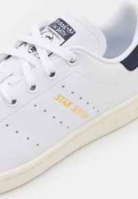 Weiße Ledersneaker mit Perforationen, marineblauen Akzenten und gelbem "STAN SMITH"-Text. Weiße Schnürsenkel und beige Gummisohle.