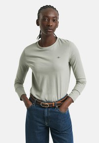 Lys grøn langærmet t-shirt med rund halsudskæring og lille broderet logo på brystet, sammen med mørkeblå jeans og et brunt bælte.