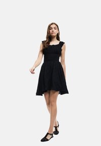 Schwarzes Kleid aus strukturiertem Stoff, tailliertes Oberteil, Rüschenträger und ausgestelltrock. Schwarze Schuhe mit T-Riemen-Design runden den Look ab.