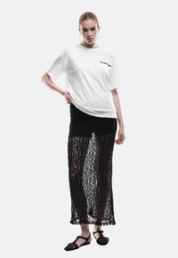 Ung kvinde med håret sat op, iført store hoop øreringe, en hvid grafisk t-shirt, et langt sort blonde skirt og sorte sandaler.