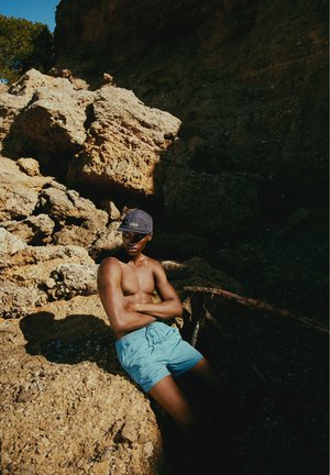 Ung mand iført blå shorts og en kasket læner sig mod store solbelyste klipper i et skyggefuldt, stenfuldt udendørs område.