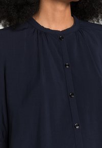 Camicia blu navy con bottoni, colletto rotondo, dettagli plissettati e bottoni neri lungo il davanti. Texture morbida e liscia.