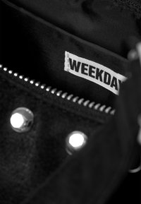 Čierna taška s semišovou textúrou, so železnými cvokmi, so zipsovým uzáverom a látkovou nášivkou s nápisom "WEEKDAY" vo vnútri.