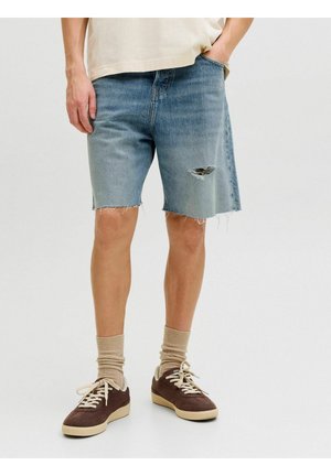 Homme portant un short en jean déchiré bleu clair, des chaussettes beiges et des baskets marron, debout devant un fond clair uni.