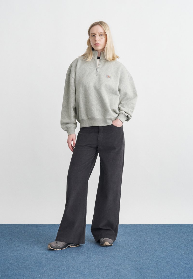 Donna con capelli biondi che indossa occhiali, felpa grigia con zip a quarto, pantaloni neri larghi e scarpe da ginnastica grigie, in piedi su pavimento blu contro parete bianca.