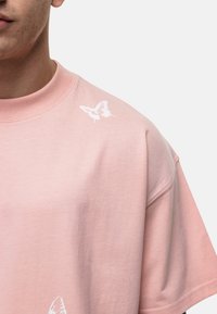 T-shirt en coton rose à manches courtes, arborant un imprimé de papillon blanc sur l'épaule gauche. Texture douce et coupe décontractée.