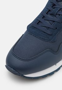 Scarpa sportiva blu navy con tomaia in rete testurizzata e pelle liscia. Presenta lacci piatti e una suola intermedia bianca a contrasto con suola esterna testurizzata.