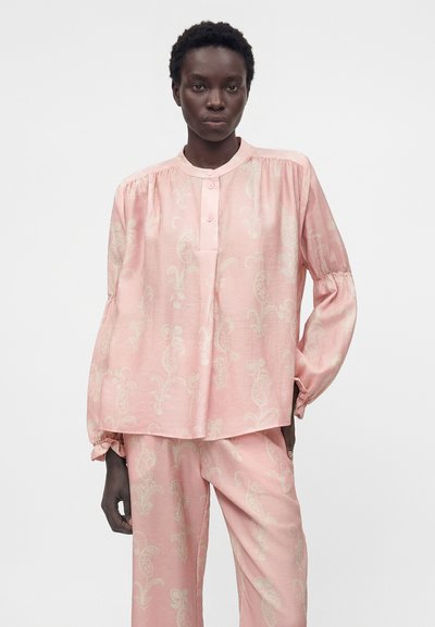 Γυναίκα που φορά φαρδιά ανοιχτό ροζ μπλούζα και ταιριαστό παντελόνι με διακριτικό σχέδιο πεϊσλί, στέκεται μπροστά σε απλό φωτεινό φόντο.