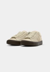 Sneaker in suede beige con lacci piatti, punta rotonda e suole in gomma marrone scuro, con un logo discreto di lato.
