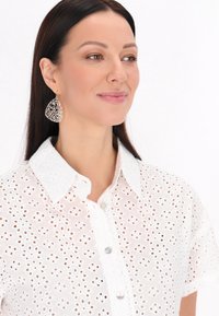 Camicia bianca in cotone con maniche corte e colletto. Presenta una chiusura frontale con bottoni e motivi intagliati intricati. Tessuto leggero e arioso.