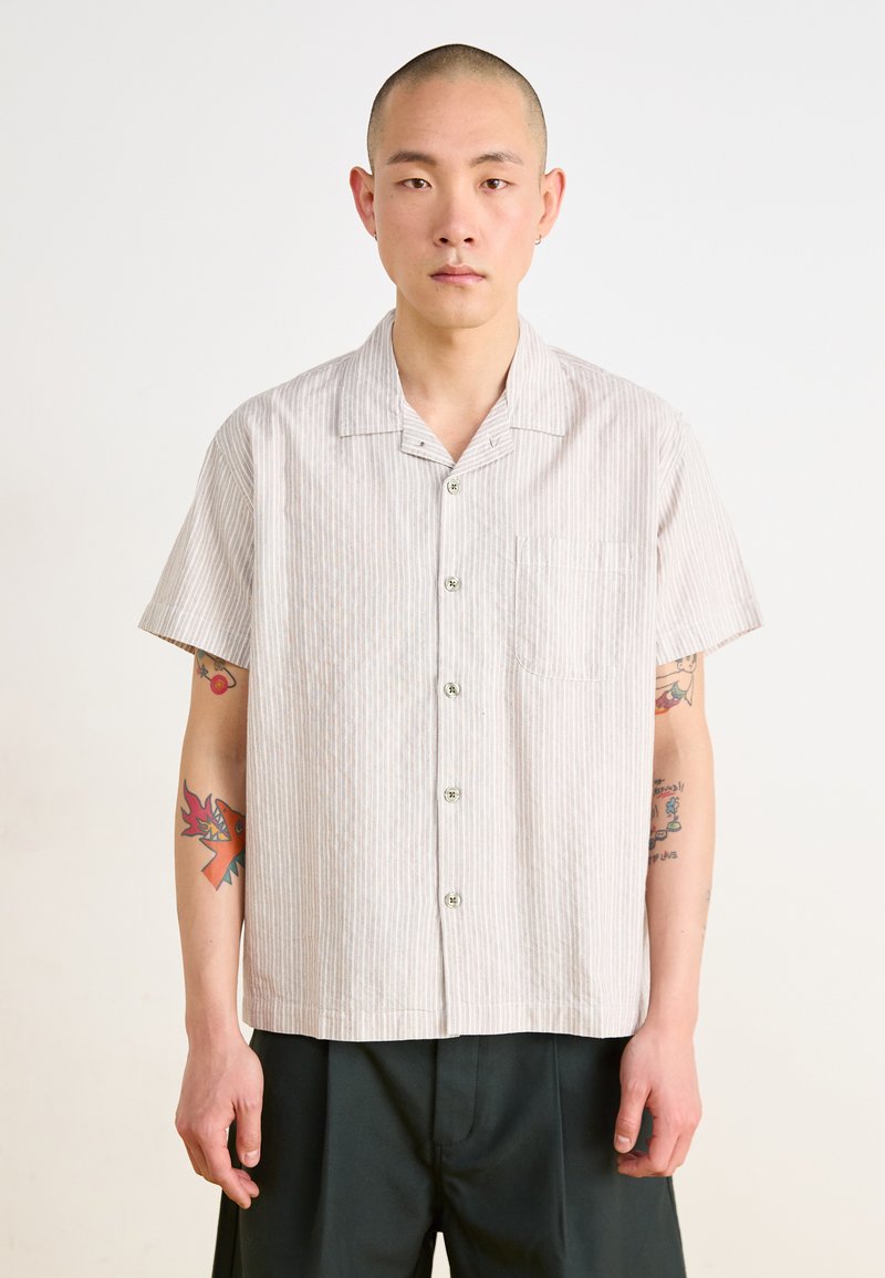Chemise à manches courtes rayée en beige clair et blanc, dotée d'un col, de six boutons et d'une poche poitrine ; tissu en coton avec une texture douce.