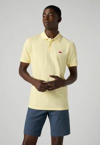 Gul pikétröja med kort krage och röd logotyp, kombinerad med mörkblå shorts. Tyget verkar slätt och något strukturerat.