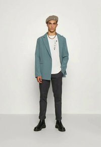 Young man wearing a beige beret, teal blazer, white graphic t-shirt, black rolled-up pants, and black boots, standing against a plain background.