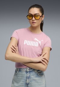 Světle růžové bavlněné tričko s bílým logem Puma. Model nosí oversized sluneční brýle v barvě jantaru. Kombinováno s denimovými šortkami. Ležérní střih.