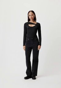 Μαύρο κορμάκι με μακριά μανίκια και λαιμόκοψη με ανοίγματα, συνδυασμένο με ψηλόμεσα μαύρα τζιν καμπάνα και μποτάκια μέχρι τον αστράγαλο, σε ουδέτερο φόντο.