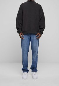 Persona que lleva una sudadera negra holgada, vaqueros azules y zapatillas blancas, de pie frente a un fondo gris liso.