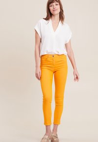 Witte, korte mouwen, V-hals top gecombineerd met fel oranje slim-fit jeans en beige loafers, tegen een neutrale achtergrond.