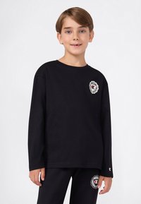 Młody chłopiec o jasnobrązowych włosach, ubrany w czarny długi rękaw bluzę oraz pasujące spodnie z białymi i czerwonymi naszywkami z logo.