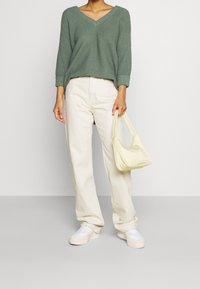 Maglione verde a maglia con scollo a V, pantaloni beige a gamba dritta, sneakers bianche e una borsa a spalla leggera di colore giallo. Sottile texture nel maglione.