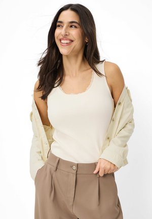 Glimlachende vrouw die een crèmekleurige tanktop met kanten rand draagt, een beige open shirt en een hoge taillebruine broek met de handen in de zakken.
