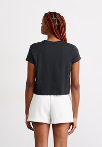 Mulher com longas tranças vermelhas, vestindo uma t-shirt preta curta e calções brancos com bainha dobrada, de pé de costas contra um fundo simples.