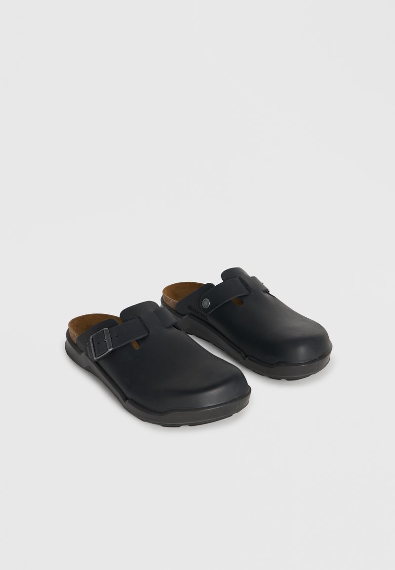 Μαύρα παπούτσια slip-on με ρυθμιζόμενους πλευρικούς ιμάντες με αγκράφες και διαμορφωμένες φτέρνες φελλού σε ουδέτερο φόντο.