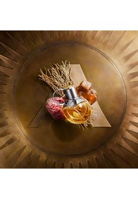 Ronde bronzen voet met zonnestraalpatroon, met een glazen parfumfles, een rood-roze bloem, karamelstukjes en kunstzinnig gerangschikte wortels.