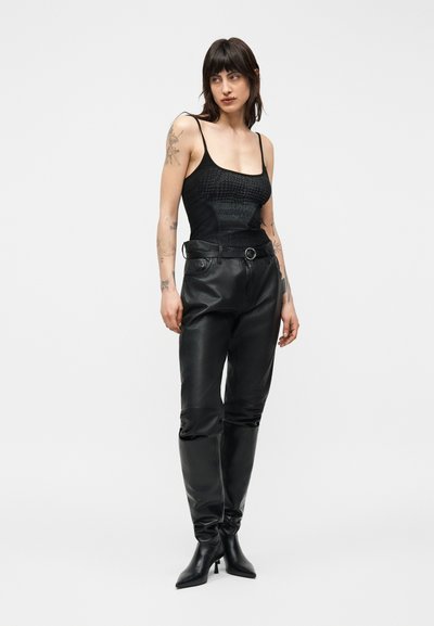 Femme tatouée portant un haut noir sans manches, un pantalon en cuir taille haute avec ceinture et des bottes noires à talons, debout devant un fond blanc.