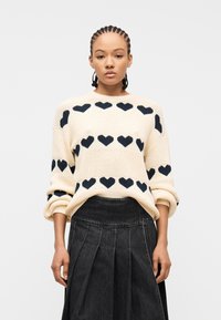 Γυναίκα με πλεξούδες φοράει κρεμ πουλόβερ με μοτίβα μαύρης καρδιάς και πλισέ σκούρα τζιν φούστα, στέκεται μπροστά σε λευκό φόντο.