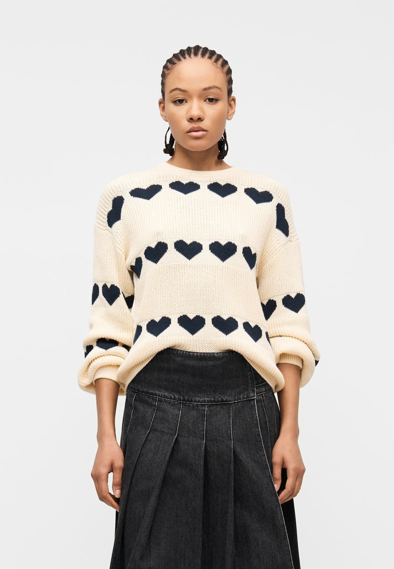 Γυναίκα με πλεξούδες φοράει κρεμ πουλόβερ με μοτίβα μαύρης καρδιάς και πλισέ σκούρα τζιν φούστα, στέκεται μπροστά σε λευκό φόντο.